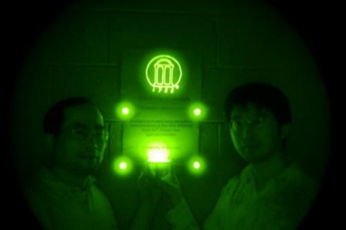 Zhengwei Pan and Feng Liu stand in a darkened room lit only by ceramic discs containing a persistent near-IR phosphor. The phosphorescent material was also mixed into the paint that was used to create the University of Georgia logo behind them. Imaging parameters: auto, ISO 200, and 3-4 seconds of exposure time using a night-vision monocular.