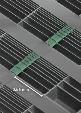 A scanning electron microscope image shows an array of optomechanical accelerometer devices with differing test-mass sizes. Highlighted green areas represent proof masses suspended by nanoscale tethers across the open, etched areas of the chip. The nanobeam cavities are seen as small square notches in the centers of the masses.