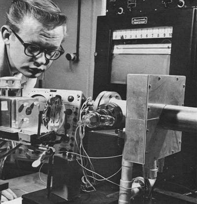 FIGURE 1. An oscilloscope mounted on a car is just below the technician&apos;s jaw and behind a lens in this photo from the Honeywell Research Center in Hopkins, MN, published in the January 1, 1965 issue. It was used to measure heating produced by a ruby laser beam focused through the large lens.