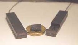 Polymer-encapsulated lasers contain rhombic Nd:YAG laser crystals (visible by the greenish reflection from their thin-film coatings). A fully encapsulated laser 50 × 10 × 3 mm in size with fiber-pump source is at left; the partly encapsulated laser at right was used in the experiments. The laser at center has no fiber-pump source. The coin seen at center as a size reference is a Swedish 10-kronor coin 20.5 mm in diameter.