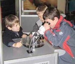 Using a microscope, children examine several samples on a turntable that have been laser microprocessed: a stent, a tooth, porous plastic foil, gummi bears, and a microscopic statue of Venus.