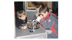 Using a microscope, children examine several samples on a turntable that have been laser microprocessed: a stent, a tooth, porous plastic foil, gummi bears, and a microscopic statue of Venus.