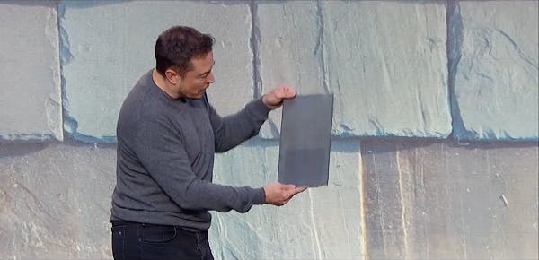 Tesla CEO Elon Musk is seen holding a tile amid an image of one style of the Tesla solar roof; note the solar panel just under the semi-transparent tile surface.