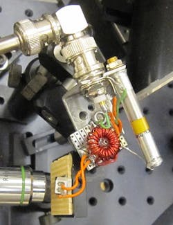 A JILA instrument generates terahertz radiation. Ultrafast pulses of near-IR laser light enter through the lens at left, striking a semiconductor wafer studded with electrodes (transparent square barely visible under the white box connected to orange wires) bathed in an oscillating electric field. The light dislodges electrons, which accelerate in the electric field and emit terahertz radiation.