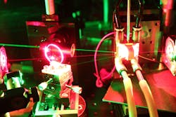 FIGURE 3. A MECSEL setup shows resonator mirrors (left and right edge of photo), intra-cavity birefringent filter for frequency stabilization and tuning (left edge), pump optics (behind left edge), the gain element (right of center) consisting of two plane-parallel diamond heat spreaders with 0.5 mm thickness each between which is sandwiched the GaInP-quantum-well gain membrane. The MECSEL 595 mW output power at 657 nm and heatsink temperature of 10°C. FIGURE 3. A MECSEL setup shows resonator mirrors (left and right edge of photo), intra-cavity birefringent filter for frequency stabilization and tuning (left edge), pump optics (behind left edge), the gain element (right of center) consisting of two plane-parallel diamond heat spreaders with 0.5 mm thickness each between which is sandwiched the GaInP-quantum-well gain membrane. The MECSEL 595 mW output power at 657 nm and heatsink temperature of 10°C.