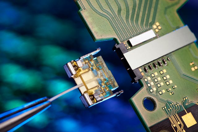 FIGURE 3. Intel&apos;s 50 Gbit/s silicon photonics transmitter and fiber connector are shown. The wide metal stripe at right is the transmitter module, bonded as a flip chip to the circuit board. The protruding metal pins align the smaller plug-in module containing the fiber, at center, which has been disconnected. The fiber extends toward lower left. Receiver is not shown.