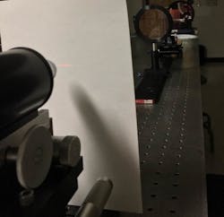 FIGURE 4. A parabolic off-axis collimator (upper right) illuminates a positive power lens with a Ronchi amplitude grating (behind lens). The array of focused spots is visible on the white screen (left) in front of the microscope objective. FIGURE 4. A parabolic off-axis collimator (upper right) illuminates a positive power lens with a Ronchi amplitude grating (behind lens). The array of focused spots is visible on the white screen (left) in front of the microscope objective.