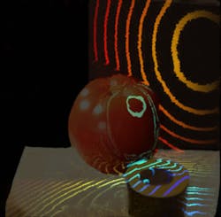 The spherical front of a light pulse illuminates a tomato and a roll of tape. The colored bands represent the pulse as it travels over time during a movie, with each band separated by approximately 20 ps. The spherical front of a light pulse illuminates a tomato and a roll of tape. The colored bands represent the pulse as it travels over time during a movie, with each band separated by approximately 20 ps.