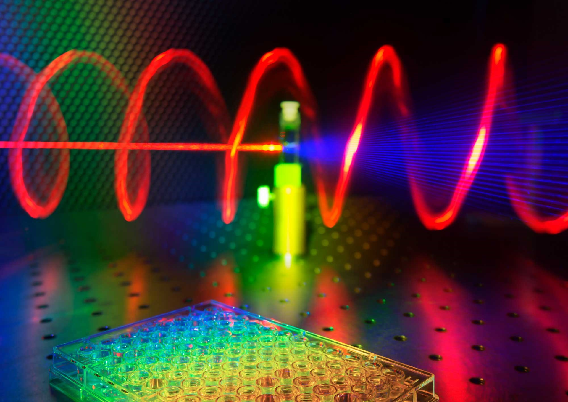 Illustration of a microplate well in the foreground, while in the background a tested sample receives red laser light and releases twisted blue light.