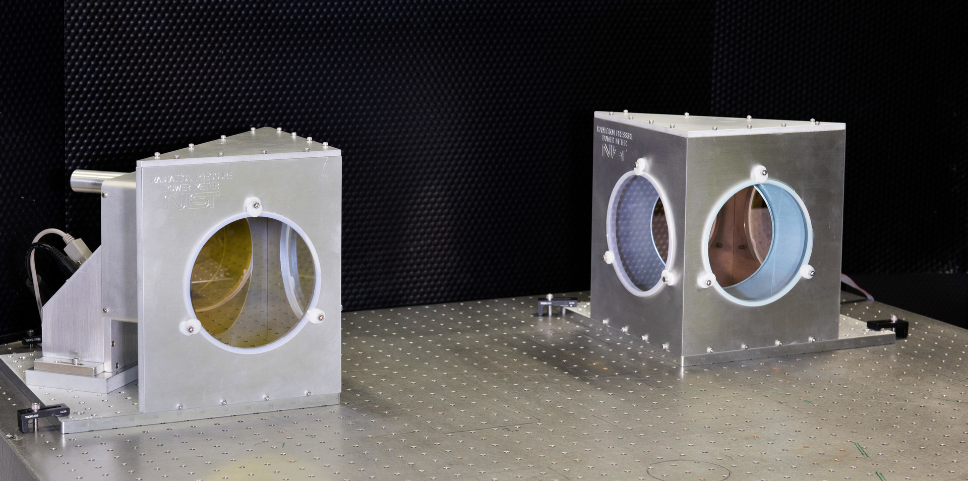 Closeup of two NIST radiation pressure power meters (RPPMs). Inside each metal housing is a mirror connected to a vertical balance. From this angle, the mirrors appear slightly yellow (left) and pink (right).