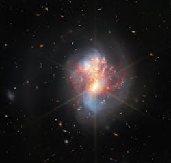 Two galaxies swirl into a single chaotic object in the center. Long blue spiral arms stretch vertically, faint at their edges. Hot gas spreads horizontally over it, mainly bright red with many small gold spots of star formation. And the core of the merging galaxies is very bright and radiates eight golden diffraction spikes. Two galaxies swirl into a single chaotic object in the center. Long blue spiral arms stretch vertically, faint at their edges. Hot gas spreads horizontally over it, mainly bright red with many small gold spots of star formation. And the core of the merging galaxies is very bright and radiates eight golden diffraction spikes.