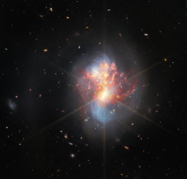 Two galaxies swirl into a single chaotic object in the center. Long blue spiral arms stretch vertically, faint at their edges. Hot gas spreads horizontally over it, mainly bright red with many small gold spots of star formation. And the core of the merging galaxies is very bright and radiates eight golden diffraction spikes.
