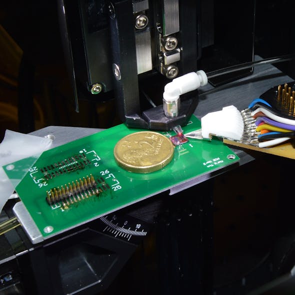 FIGURE 1. An integrated photonic chip is wire-bonded to a printed circuit board (PCB) with an Australian two-dollar coin placed for scale reference. The micron-sized metal wires are for DC testing. The chip, fabricated at an international foundry, contains micron-sized modulators, photodetectors, and heaters. It&rsquo;s made of silicon and used for microwave signal processing and telecommunications signal filtering.